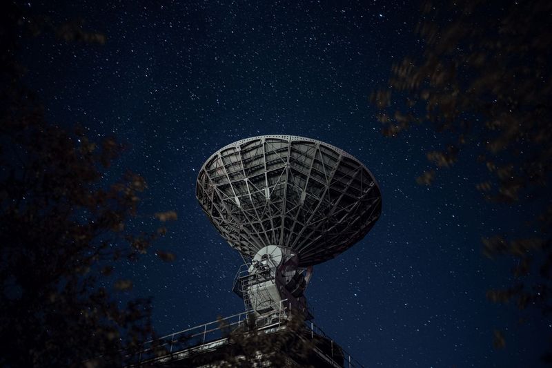From below of blue starry sky over radio telescope and trees with leaves